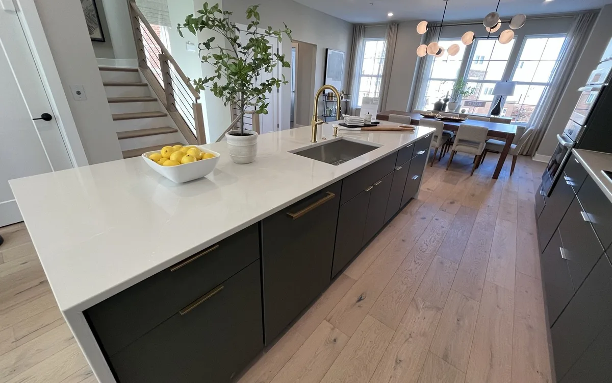 Open-plan main level renovation in a DMV townhouse showing a dark charcoal flat-panel kitchen island with white waterfall quartz countertop, brass hardware and faucet, wide-plank natural oak hardwood flooring extending into the dining area, and a modern multi-globe pendant light fixture over the dining table