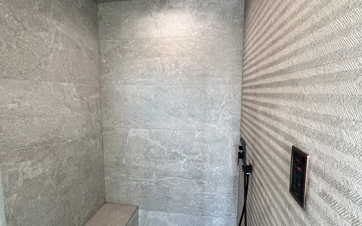 Steam shower interior with gray marble-look large-format porcelain tile walls and ceiling, black square rainfall showerhead, built-in bench, and digital steam control panel