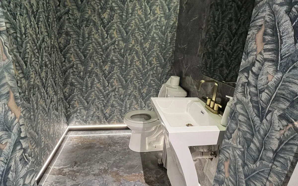 Bold tropical leaf wallpaper covering all walls of a renovated powder room with marble-look tile floor and gold fixtures