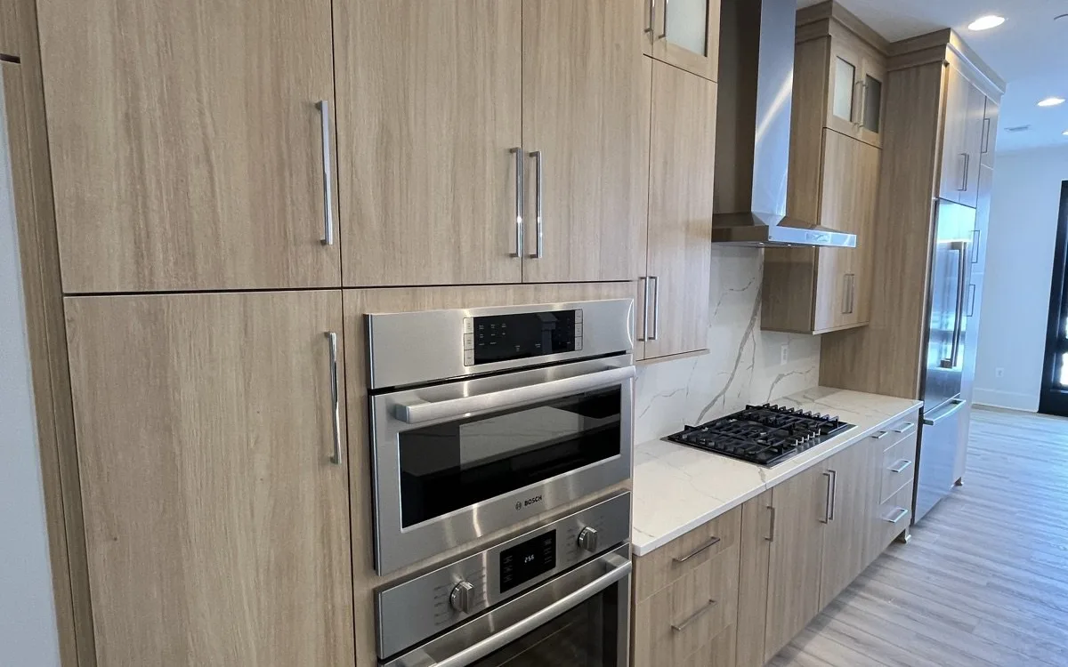 Modern kitchen renovation with floor-to-ceiling light natural wood-tone flat-panel cabinets, stacked Bosch stainless double wall oven and microwave in a tall pantry column, separate gas cooktop with calacatta marble-look porcelain slab backsplash, and light LVP flooring